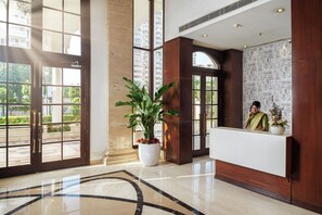 A reception area with a large potted plant, a reception desk, and a view of a building through large glass doors. - DLF Club 5 (Gurugram)