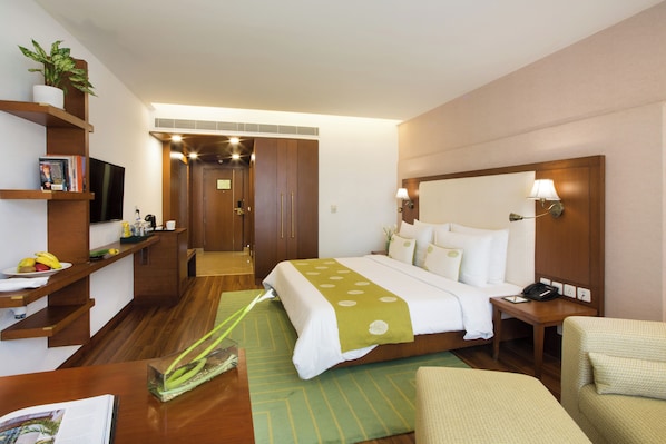 A hotel room with a bed, bedside table, lamp, chair, TV, and a small plant. - DLF Club 5 (Gurugram)