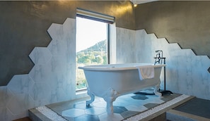 Une salle de bain moderne avec une baignoire sur pieds, des murs ornés de motifs hexagonaux et une vue sur une colline, visible à travers une grande fenêtre.