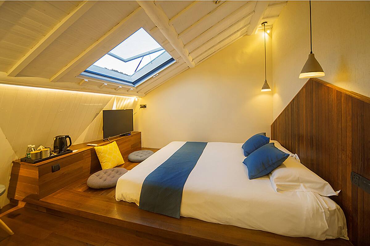 A bedroom with a skylight, a bed with blue and white bedding, a bedside table with a lamp, and a TV.