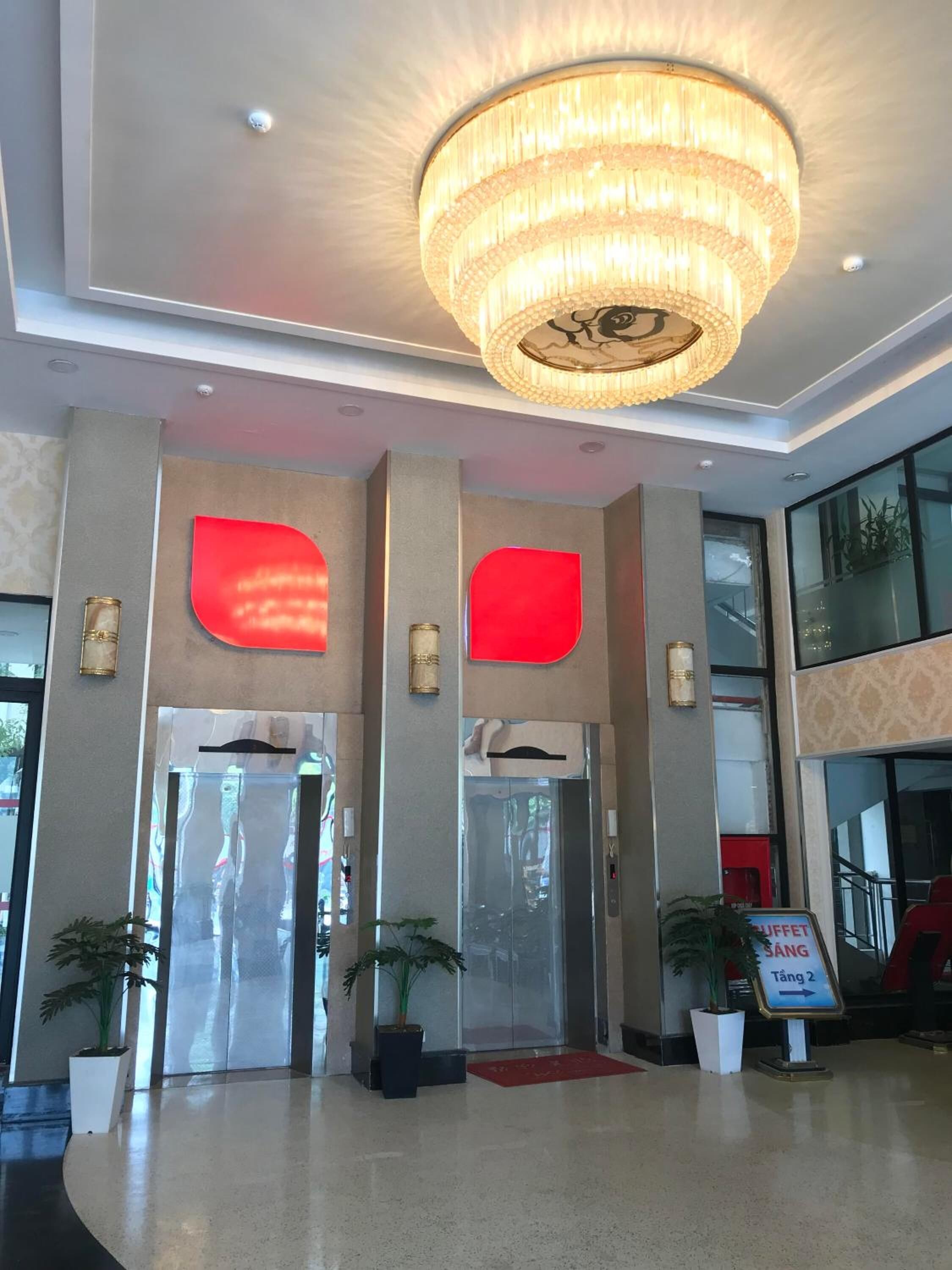 Eine Hotelhalle mit einer Chandelüstere, Aufzügen und einem Schild, das den Weg zum Frühstücksbuffet zeigt.