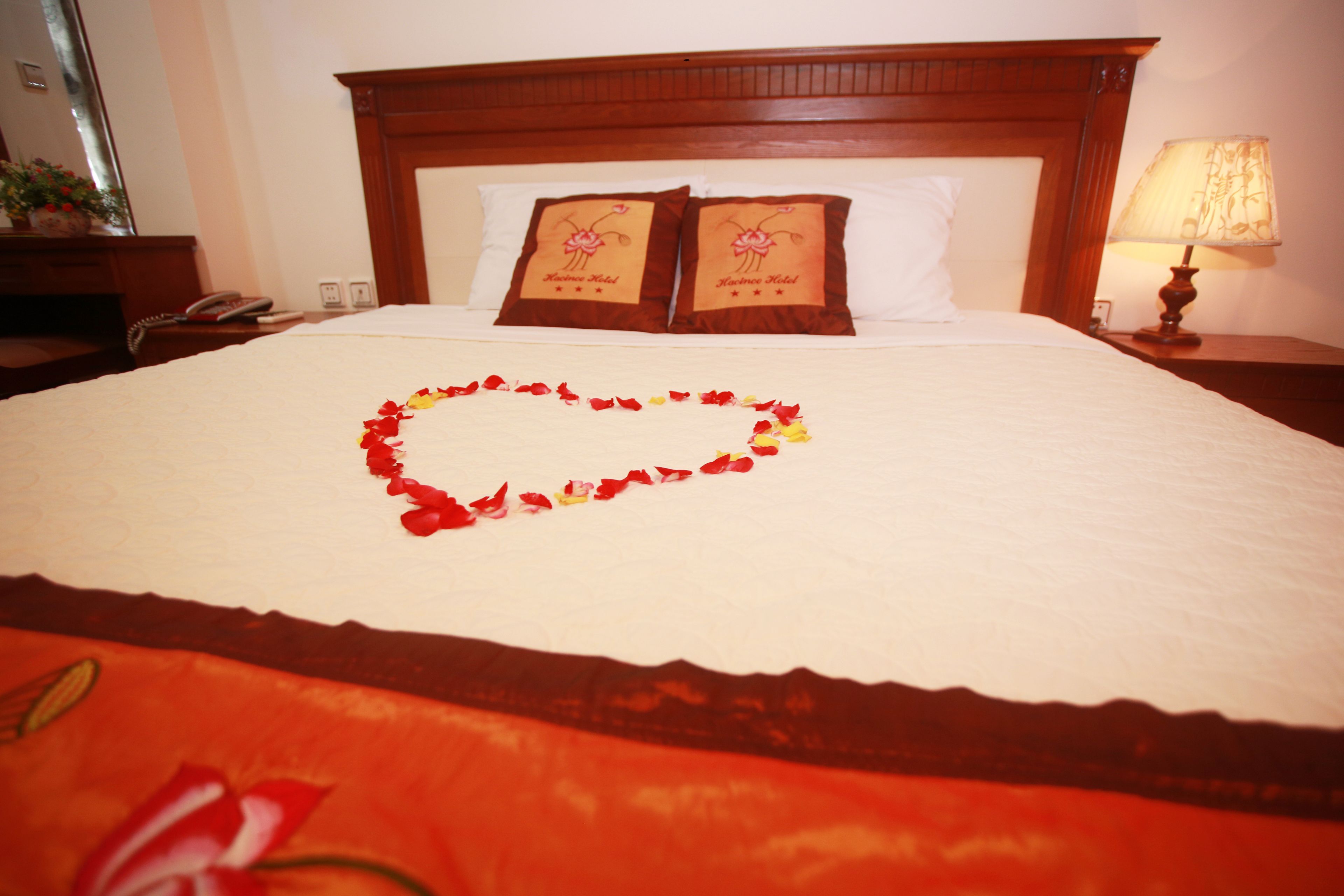 A hotel room with a neatly made bed, a bedside lamp, and a floral heart decoration on the bed.