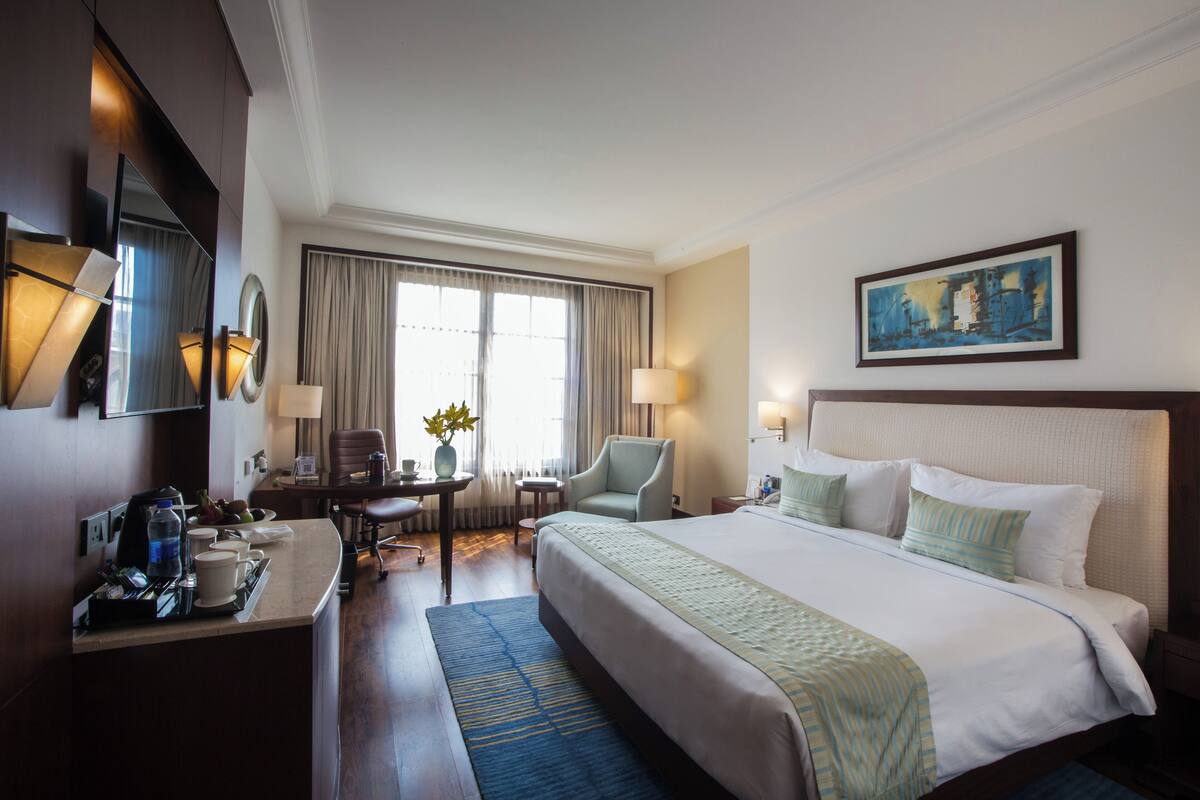 A hotel room with a large bed, a desk with a lamp, a chair, and a painting on the wall.