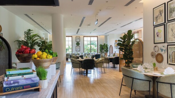 A modern dining area with a long counter, multiple tables, and chairs. There are potted plants and a variety of books on the counter. - DLF Club 3 (Gurugram)