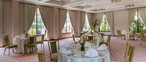 A spacious dining area with multiple round tables set for a meal, large windows with curtains, and a tiled floor.