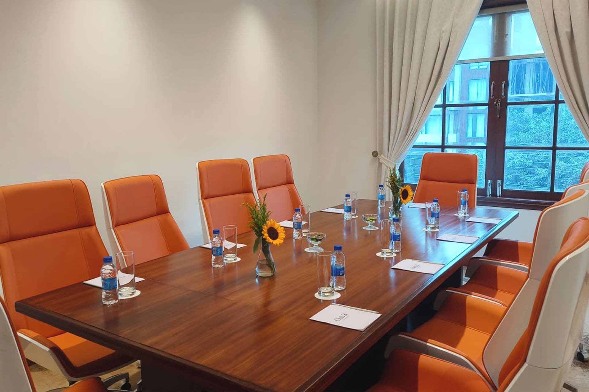 A conference room with a long table, orange chairs, water bottles, and a vase with sunflowers.