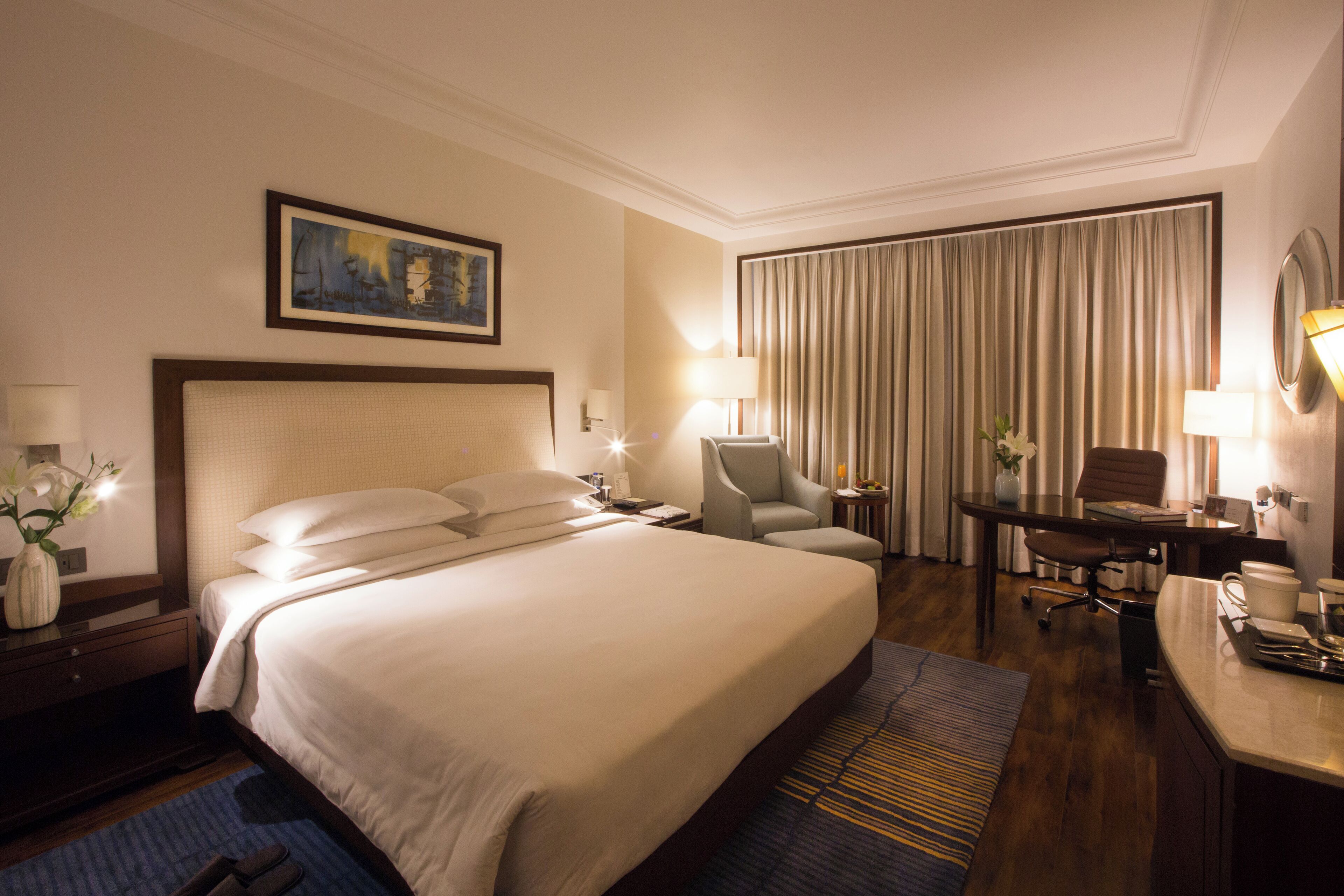 A hotel room with a large bed, a desk, a chair, a lamp, and a painting on the wall.