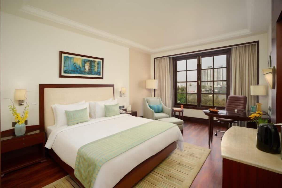 A hotel room with a large bed, two chairs, a desk, a window with curtains, and a painting on the wall.