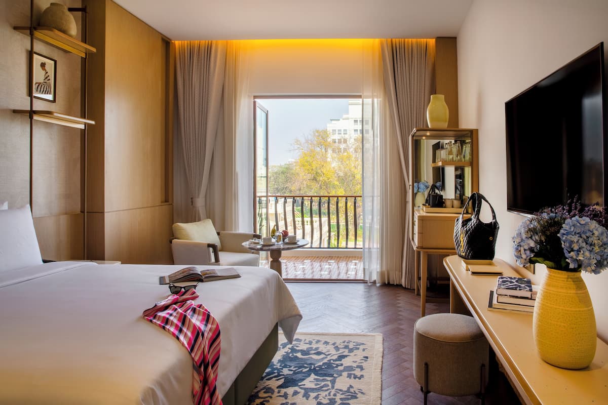Habitación de hotel con cama, escritorio, silla, televisor, ventana con cortinas y balcón con barandilla.