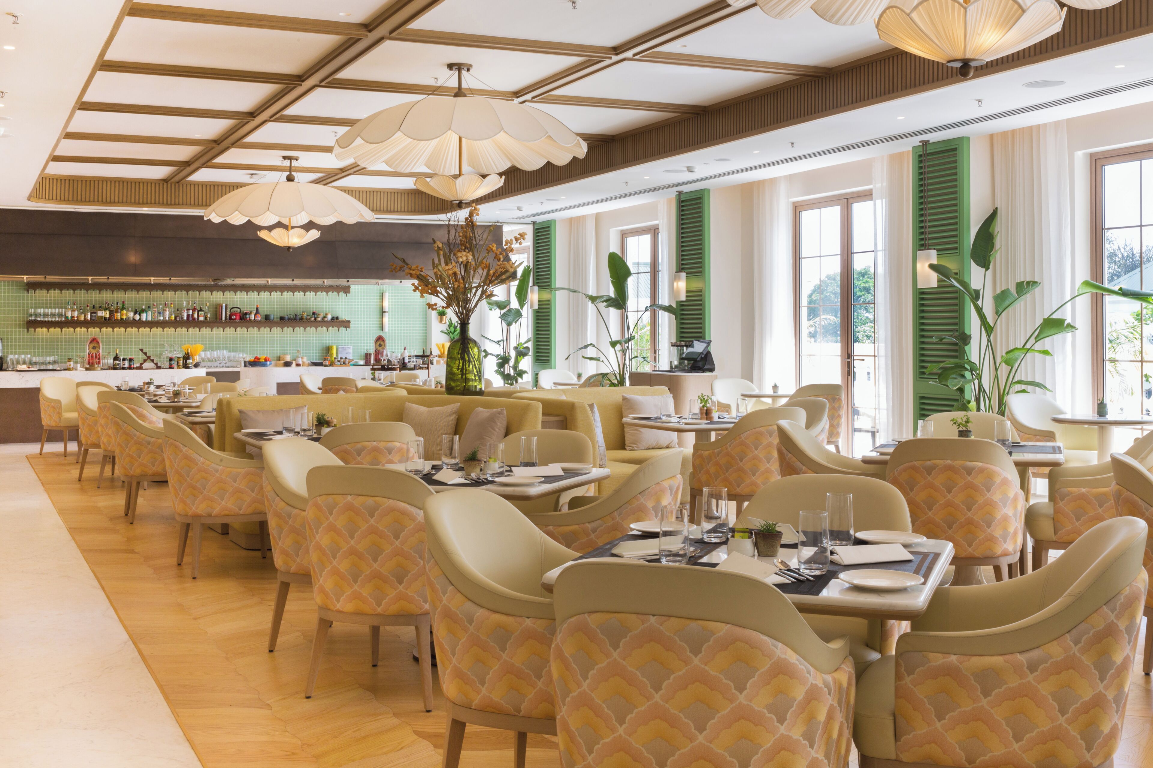 A dining area with tables and chairs, a bar in the background, and large windows with green shutters.
