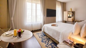 Habitación de hotel con cama, televisión, mesita de noche con un libro y un teléfono, una pequeña mesa con fruta y bebidas, y ventana con cortinas.