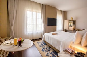 Hotelkamer met een bed, een televisie, een nachtkastje met een boek en telefoon, een kleine tafel met fruit en drankjes, en een raam met gordijnen.