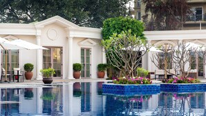 Una piscina con agua cristalina, rodeada de plantas y flores en macetas, y un edificio de estilo arquitectónico clásico.