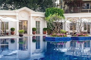 Una piscina con agua cristalina, rodeada de plantas y flores en macetas, y un edificio de estilo arquitectónico clásico.