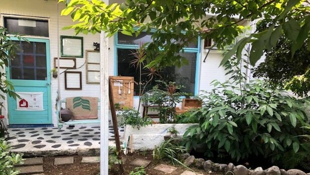 Um jardim com um caminho de pedra que leva a uma porta, rodeado por diversas plantas e um pequeno banco de madeira.