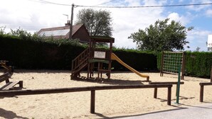 Un parque infantil con una estructura de juego de madera, que incluye un tobogán y una zona de escalada, sobre suelo arenoso, con un seto y árboles al fondo.