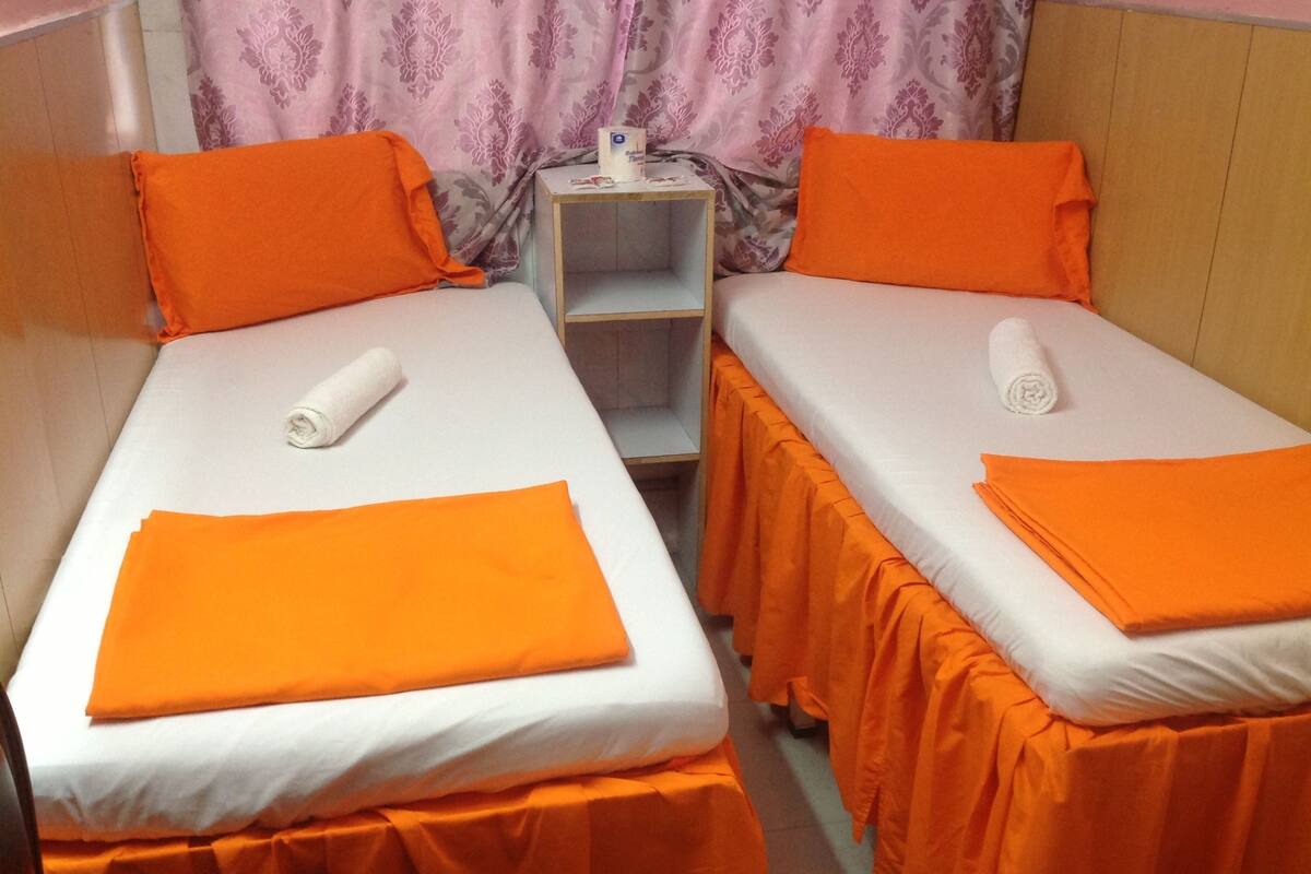 Deux lits simples avec une literie et des oreillers orange, une petite table de chevet avec une lampe et un rideau à motifs floraux.
