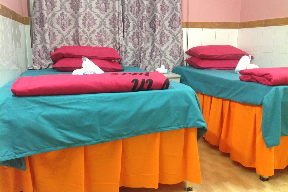 Deux tables de massage recouvertes de draps turquoise et orange, agrémentées d’oreillers rouges et d’un rideau à motifs en arrière-plan.