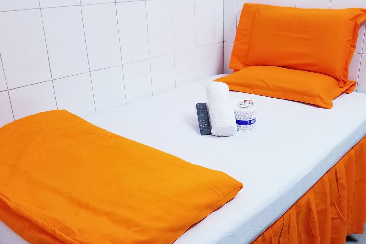 Un lit simple avec une couvre-lit orange et des coussins assortis, une télécommande et une serviette enroulée sur un matelas blanc.