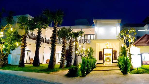 Une entrée d’hôtel bien éclairée, avec une porte centrale, des palmiers et un chemin menant à l’entrée.