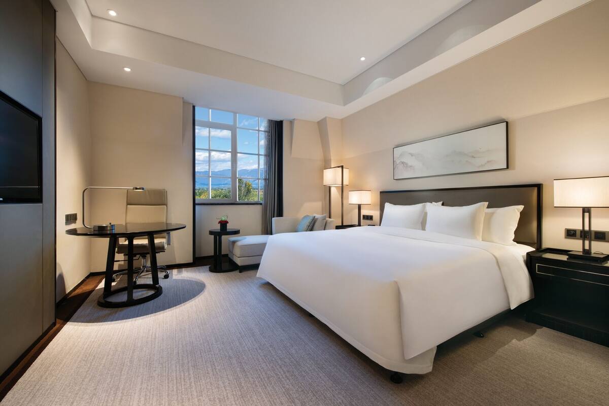 A modern hotel room with a large bed, a desk, a chair, a small table, and a window with a mountain view.
