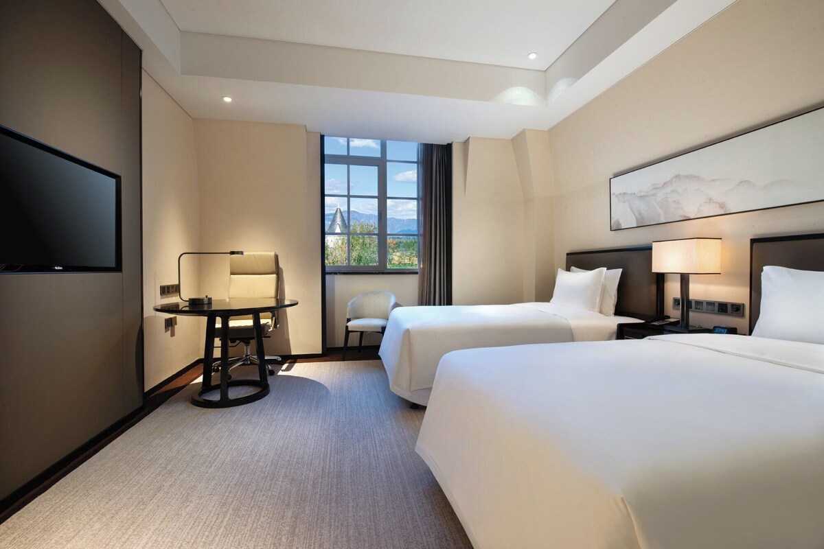Habitación de hotel con dos camas, un escritorio, una silla, un televisor y una ventana con vistas.