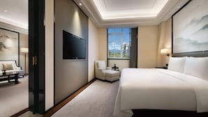 Habitación de hotel con una cama grande, un televisor de pantalla plana en la pared, una zona de estar con sofá y sillón, y vistas al exterior a través de un amplio ventanal.
