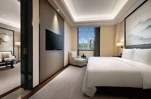 Habitación de hotel con una cama grande, un televisor de pantalla plana en la pared, una zona de estar con sofá y sillón, y vistas al exterior a través de un amplio ventanal.