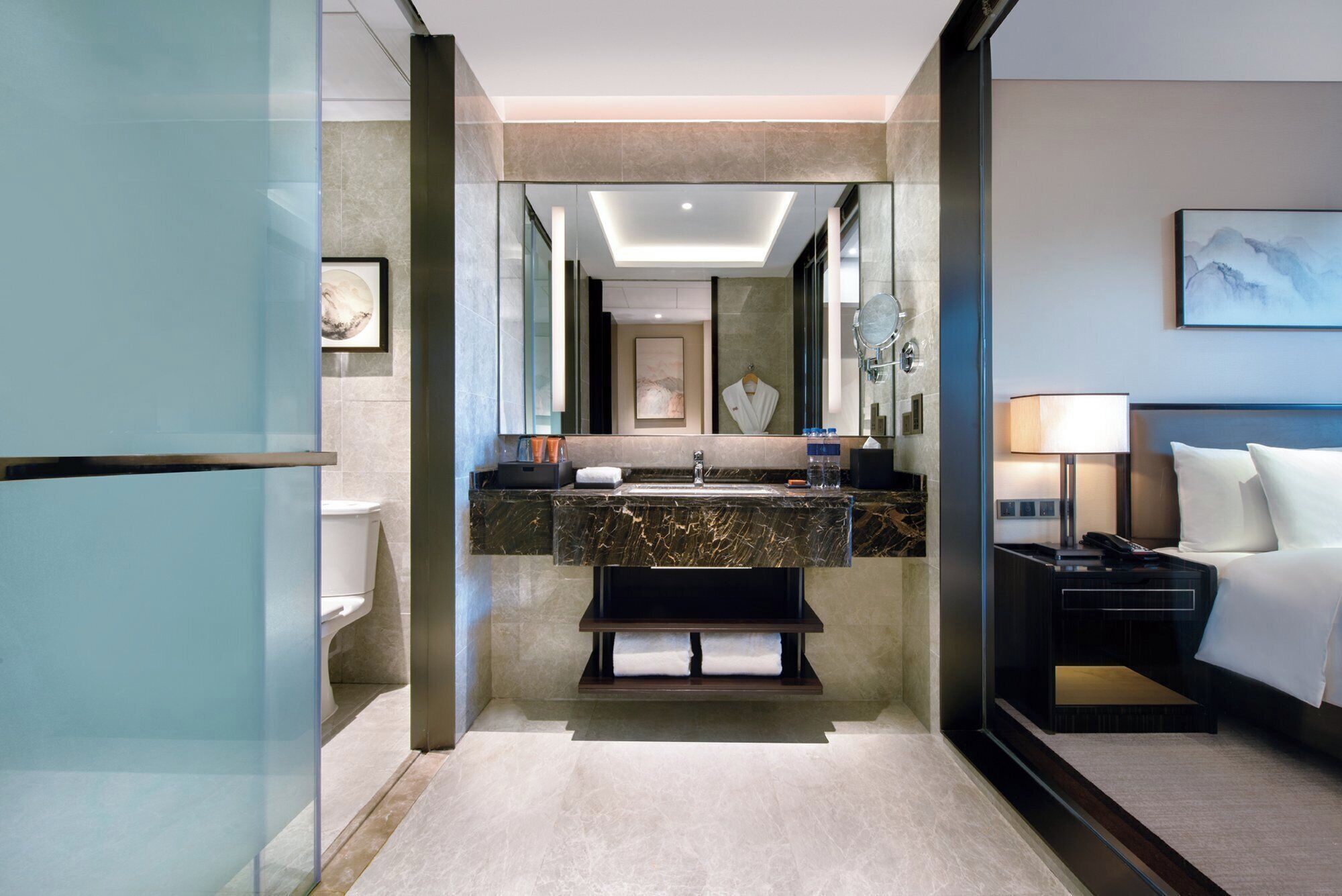 Un baño de hotel moderno con un lavamanos de mármol, un espejo grande, un inodoro y una cama con una mesita de noche.