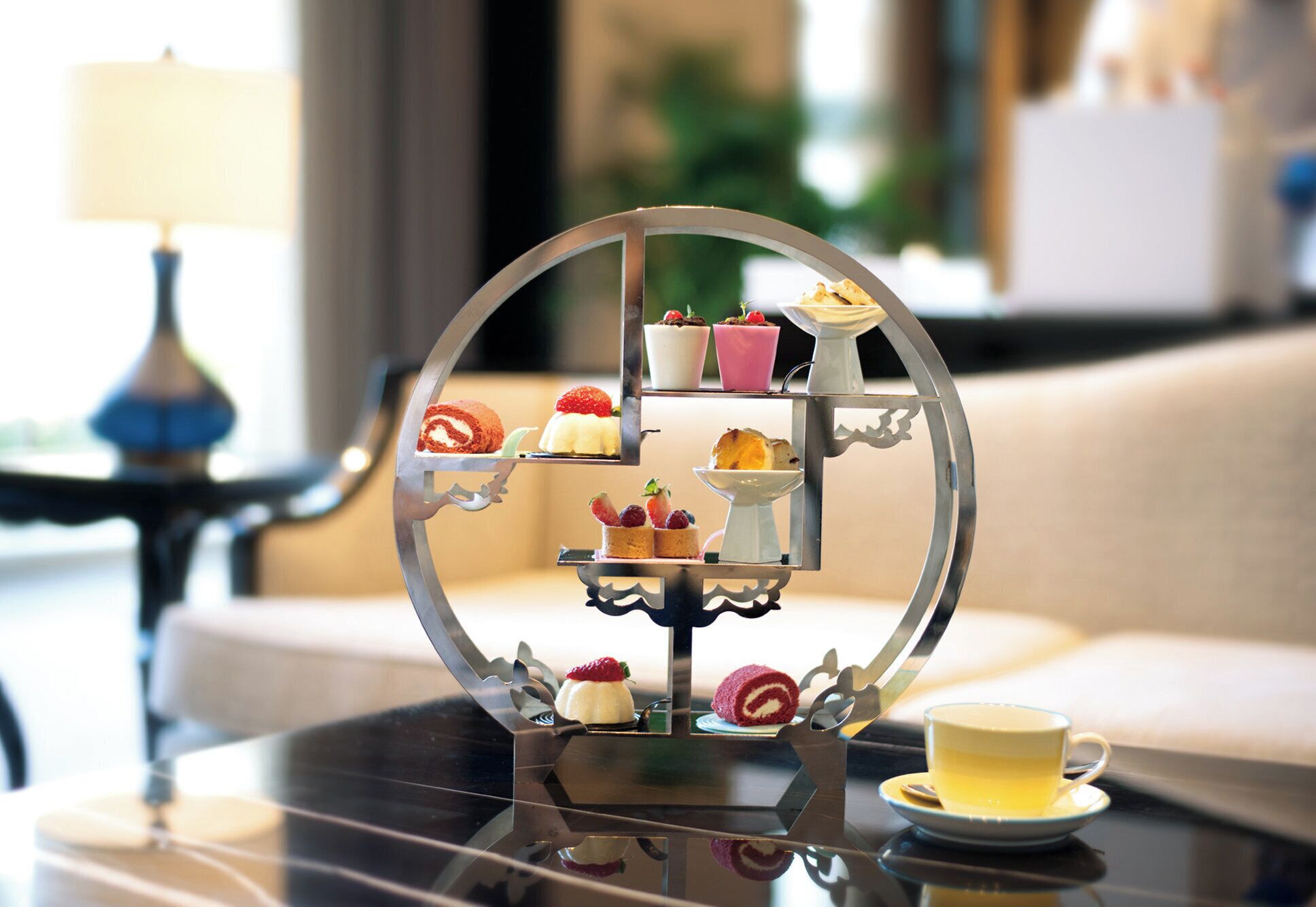 A tiered dessert stand with various pastries on a reflective surface, a cup of tea, and a lamp in the background.