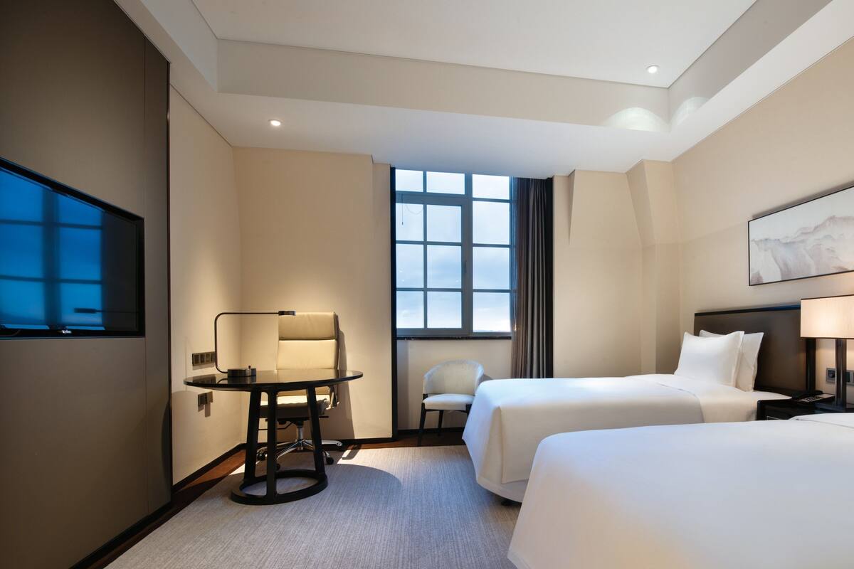 A hotel room with two beds, a desk, a chair, and a large window.