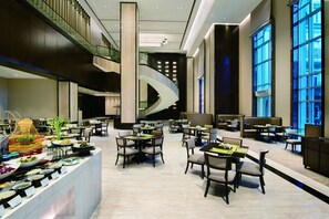 Un amplio vestíbulo de hotel con varias zonas de comedor, una escalera central y grandes ventanales.
