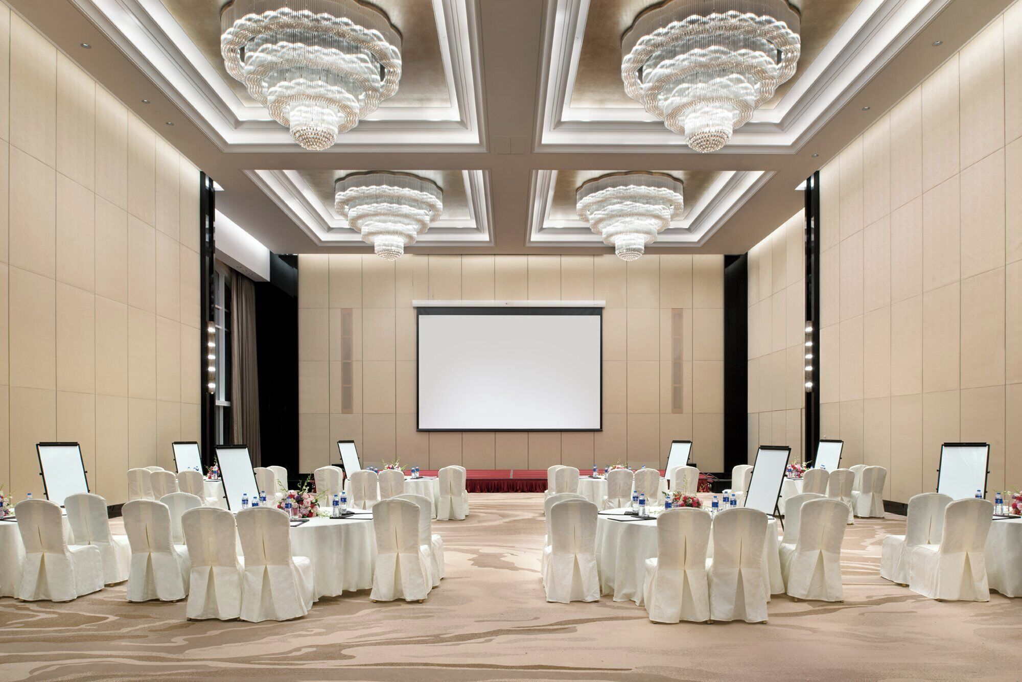 Un salón de conferencias con una pantalla grande, sillas blancas y candelabros.