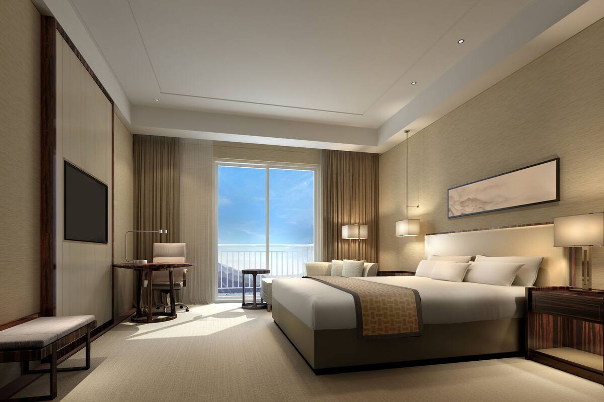 A modern hotel room with a large bed, a desk, and a chair. There is a window with a view of the sky and a balcony.