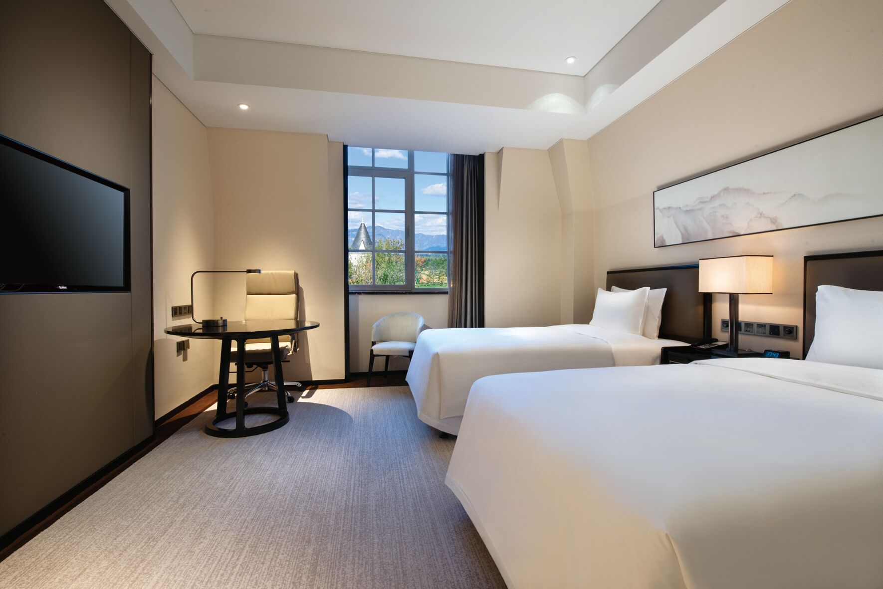 A hotel room with two beds, a desk, a chair, a TV, and a window with a view of mountains.