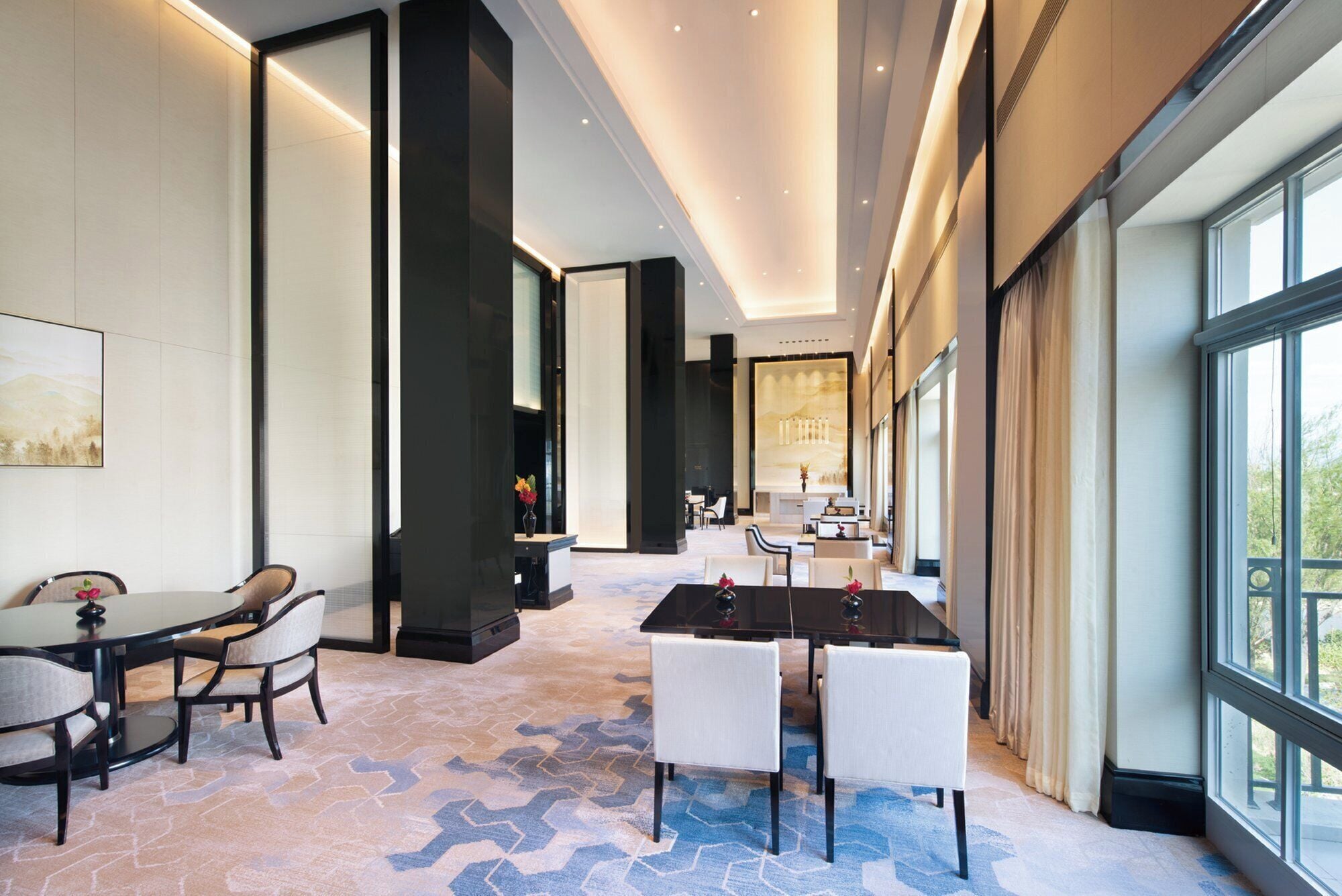 A modern hotel lobby with a large dining table, comfortable seating, and a view through large windows.