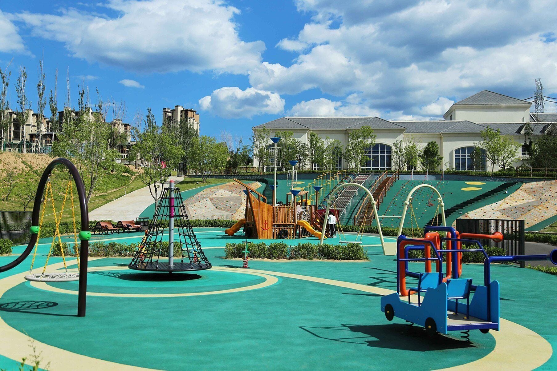 A playground with various play structures, a circular swing set, and a building in the background.
