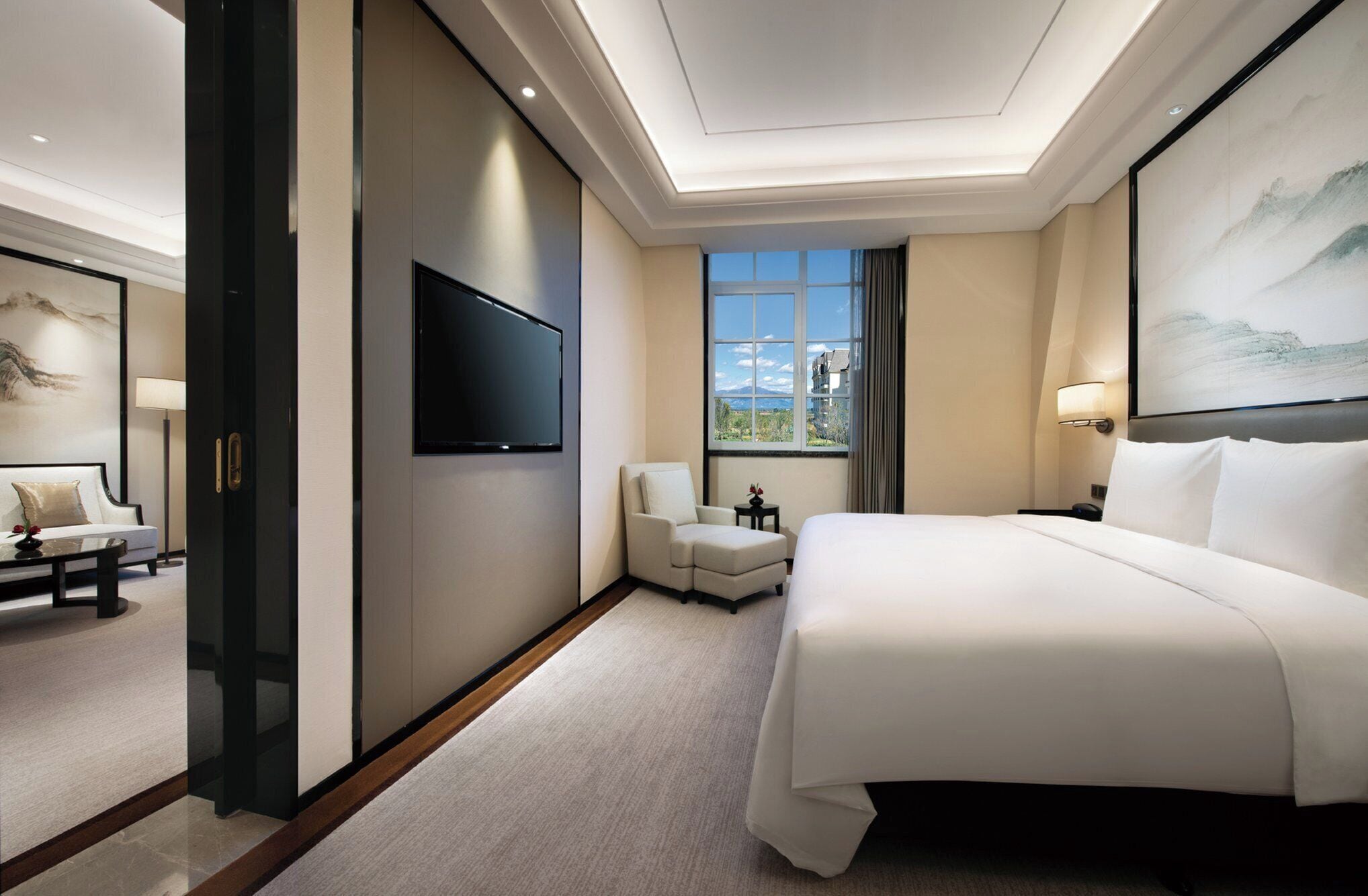 Quarto de hotel com uma cama grande, televisão de tela plana na parede, área de estar com sofá e poltrona, e janela com vista para o exterior.