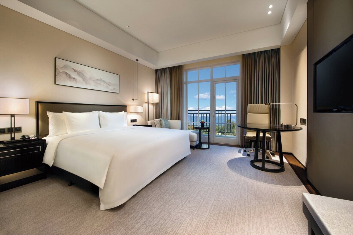 Habitación de hotel con una cama grande, un escritorio, una silla, un balcón con vistas y un televisor.