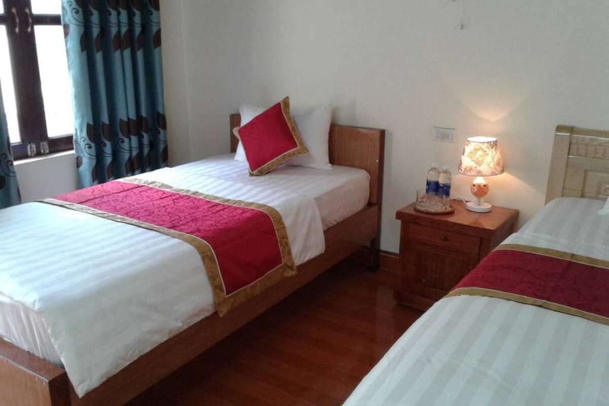 Une chambre d’hôtel avec deux lits simples, des têtes de lit en bois, une table de chevet avec une lampe et une fenêtre avec des rideaux.