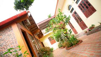 A courtyard with a brick floor, a tree with green fruit, and a building with red doors and windows.