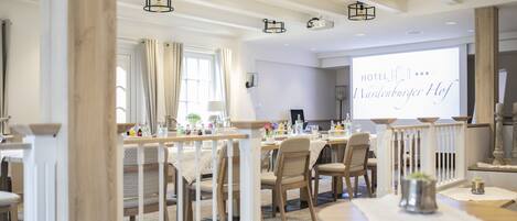 Un comedor de hotel con mobiliario de madera, barandillas blancas y una pantalla de proyección que muestra el nombre del hotel.