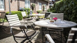 Een terras buiten met ronde tafels en stoelen, een bakstenen gebouw op de achtergrond en een haag aan de rechterkant.