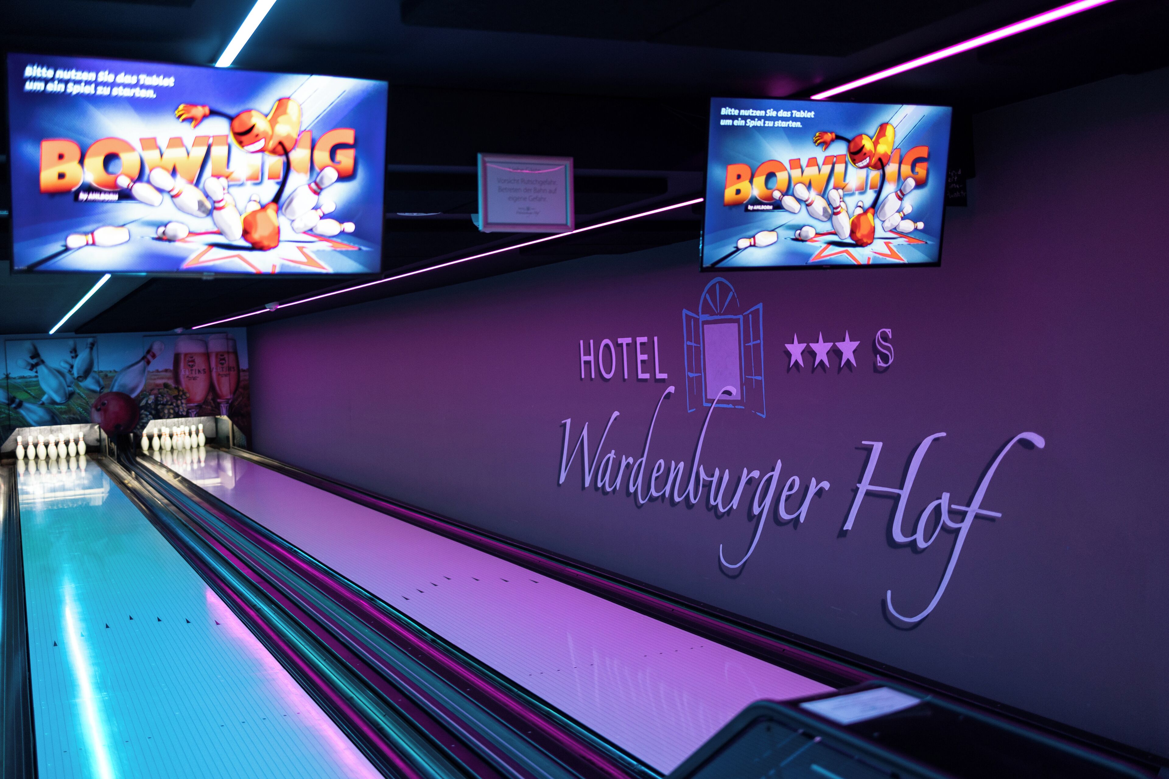 Uma pista de boliche com letreiros de neon e uma parede exibindo o nome 'Hotel Wardenburger Hof'.