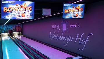 Uma pista de boliche com letreiros de neon e uma parede exibindo o nome 'Hotel Wardenburger Hof'.