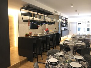 A modern kitchen with a central island, bar seating, and a dining area with round tables set for a meal. - MyHome 75 (Pescara)