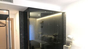 Un dormitorio moderno con una puerta de cristal que da al baño, una cama con colcha negra y gris, y una mesita de noche con una lámpara.