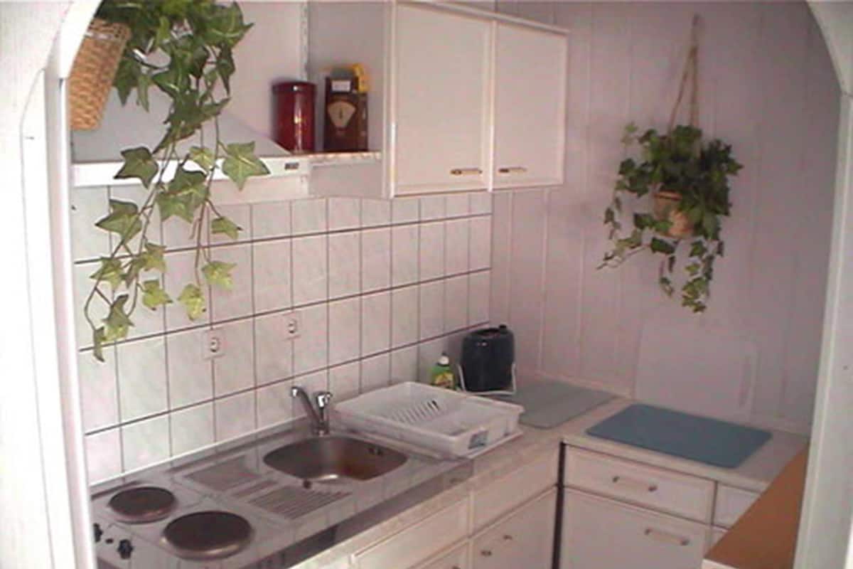 A small kitchen with white cabinets, a sink, and a stove. There are plants hanging from the ceiling and a shelf with a clock and a small appliance.