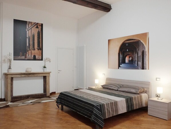 A bedroom with a large bed, a nightstand, a bedside table, a fireplace, and two wall-mounted lamps. - Centro Storico Suites (Ferrara)