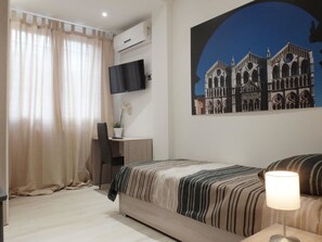 A bedroom with a bed, a desk with a chair, a TV mounted on the wall, and a window with curtains. - Centro Storico Suites (Ferrara)